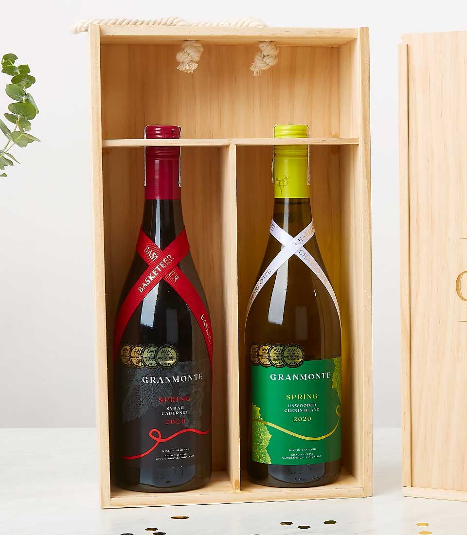A luxurious wooden box elegantly divided into two compartments, cradling two fine bottles of wine side by side. The left bottle boasts a sophisticated red label adorned with a graceful ribbon at the neck, inscribed with 
