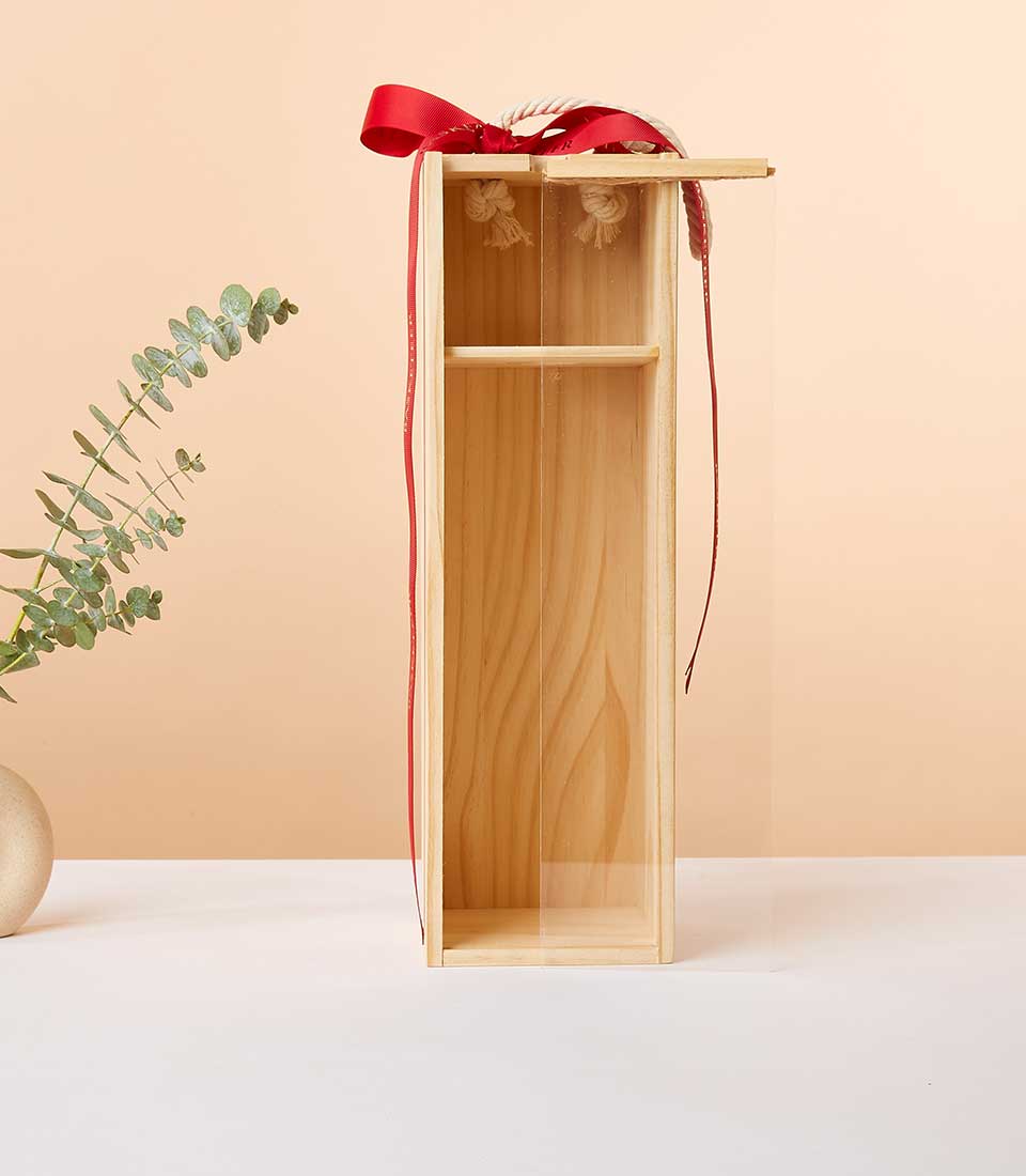 A luxurious vertical wooden gift box with an open front and a transparent panel showcases its exquisite contents. The box is adorned with a sophisticated red ribbon elegantly tied on top, featuring the Basketeer logo for an added touch of prestige. It is set against a soft beige backdrop, complemented by a minimalist vase holding fresh eucalyptus leaves to the left side, creating an inviting and refined presentation that promises indulgence.