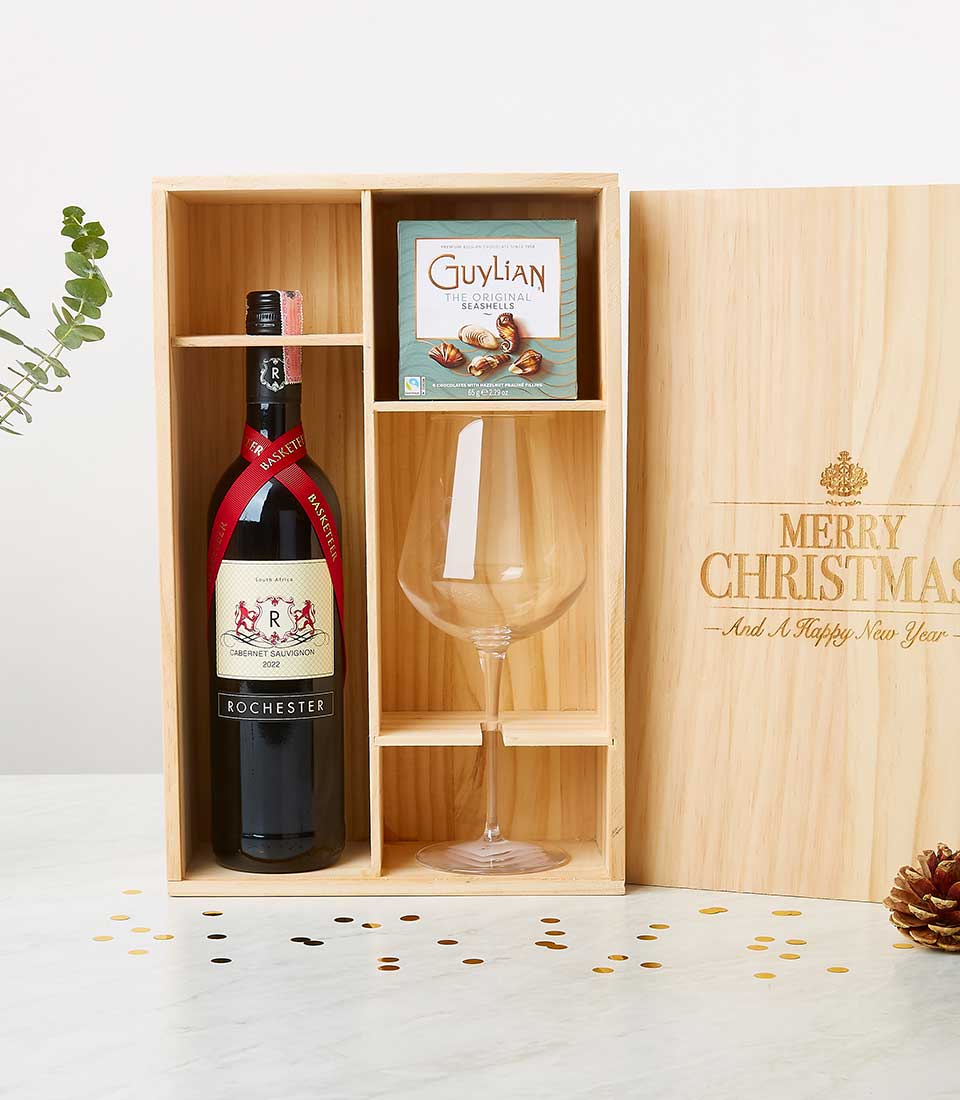 A luxurious wooden gift box from Basketeer elegantly displayed on a pristine white surface. Inside, the box reveals a bottle of Rochester wine adorned with a sophisticated red ribbon, alongside an exquisite wine glass and an opulent box of Guylian chocolates. The lid, gracefully resting to the right, is beautifully engraved in gold with 