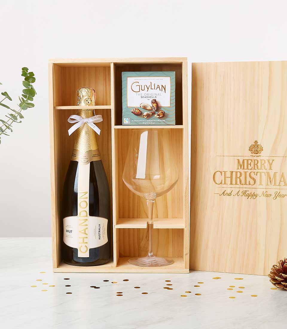 An opulent holiday gift set exudes elegance with a bottle of Chandon Brut, a large crystal wine glass, and an exquisite box of Guylian chocolates, all meticulously arranged in a finely crafted wooden box. The lid of the box is beautifully inscribed with 
