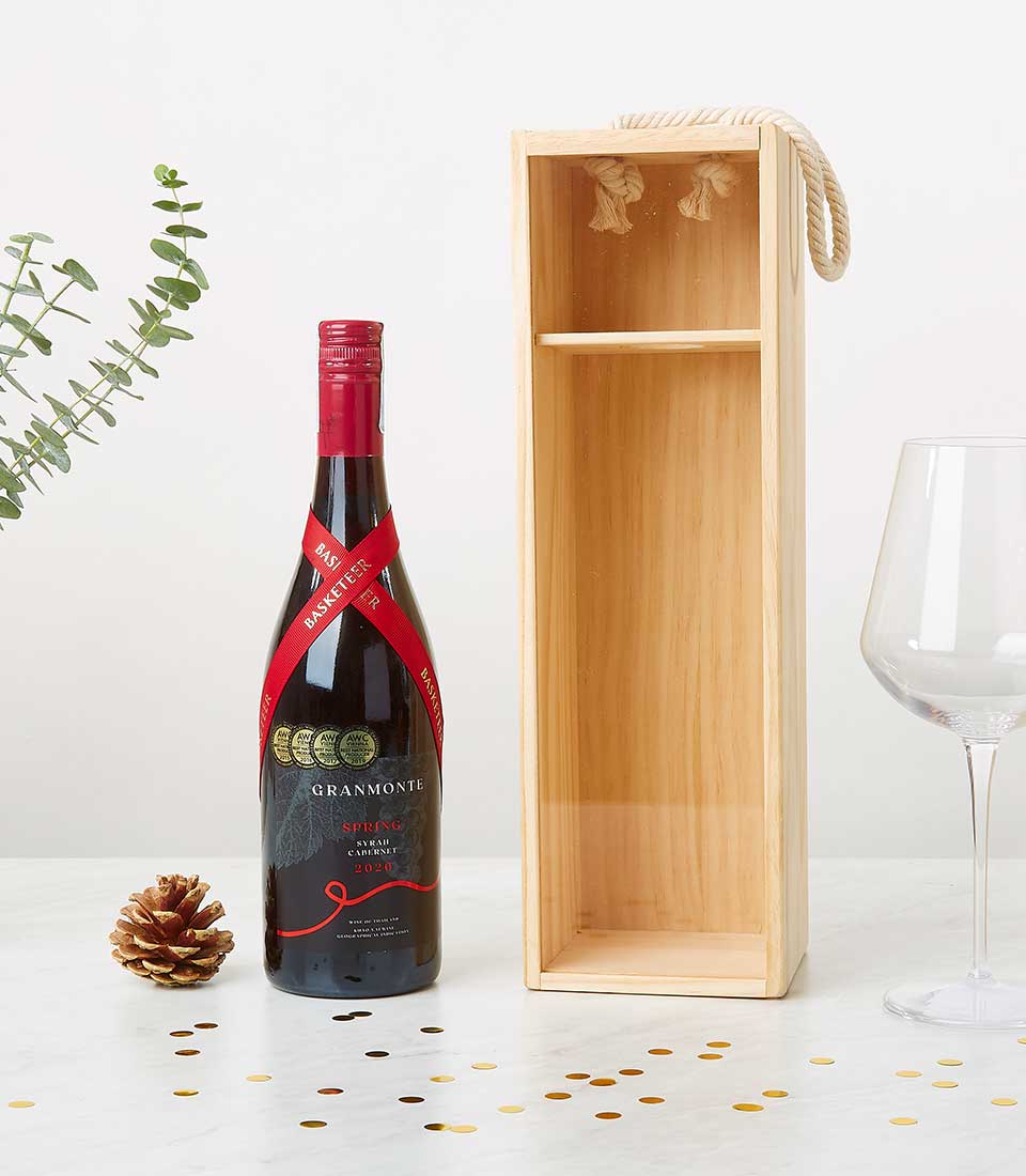 A sumptuous bottle of red wine, elegantly adorned with a crimson ribbon, rests beside an exquisite wooden wine box featuring a rope handle and distinguished Basketeer branding. A delicate wine glass stands ready to savor the moment, surrounded by festive pine cones and shimmering confetti. Lush green leaves provide a serene backdrop, all gracefully arranged on a pristine white surface to create an atmosphere of luxurious indulgence.