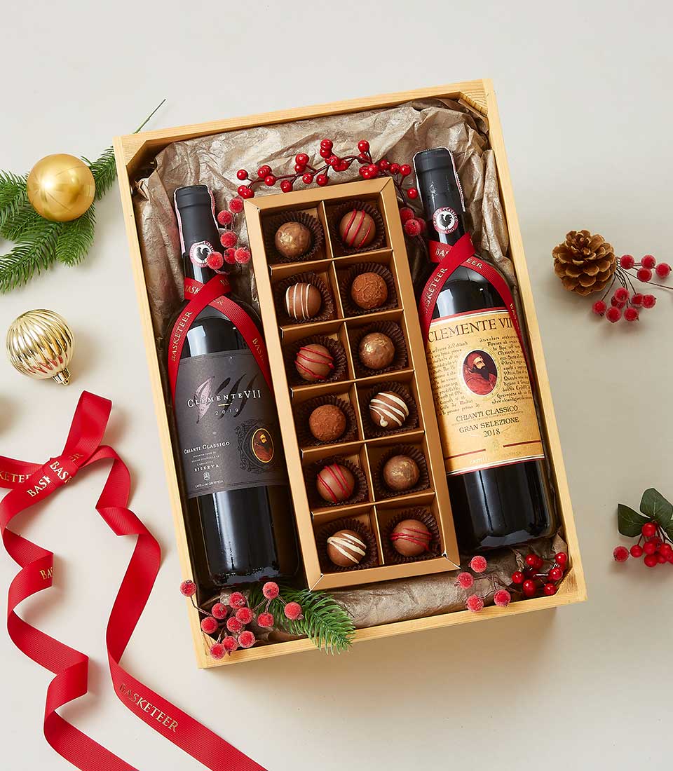 A luxurious wooden gift box elegantly cradles two exquisite bottles of red wine on the left and right, with a central tray brimming with an assortment of gourmet chocolates. The scene is adorned with festive decorations such as vibrant red ribbons, glistening red berries, a shimmering gold ornament, and lush pine branches. The Basketeer logo graces the velvet ribbon encircling the box, emphasizing the opulence and thoughtfulness of this perfectly curated gift ensemble.