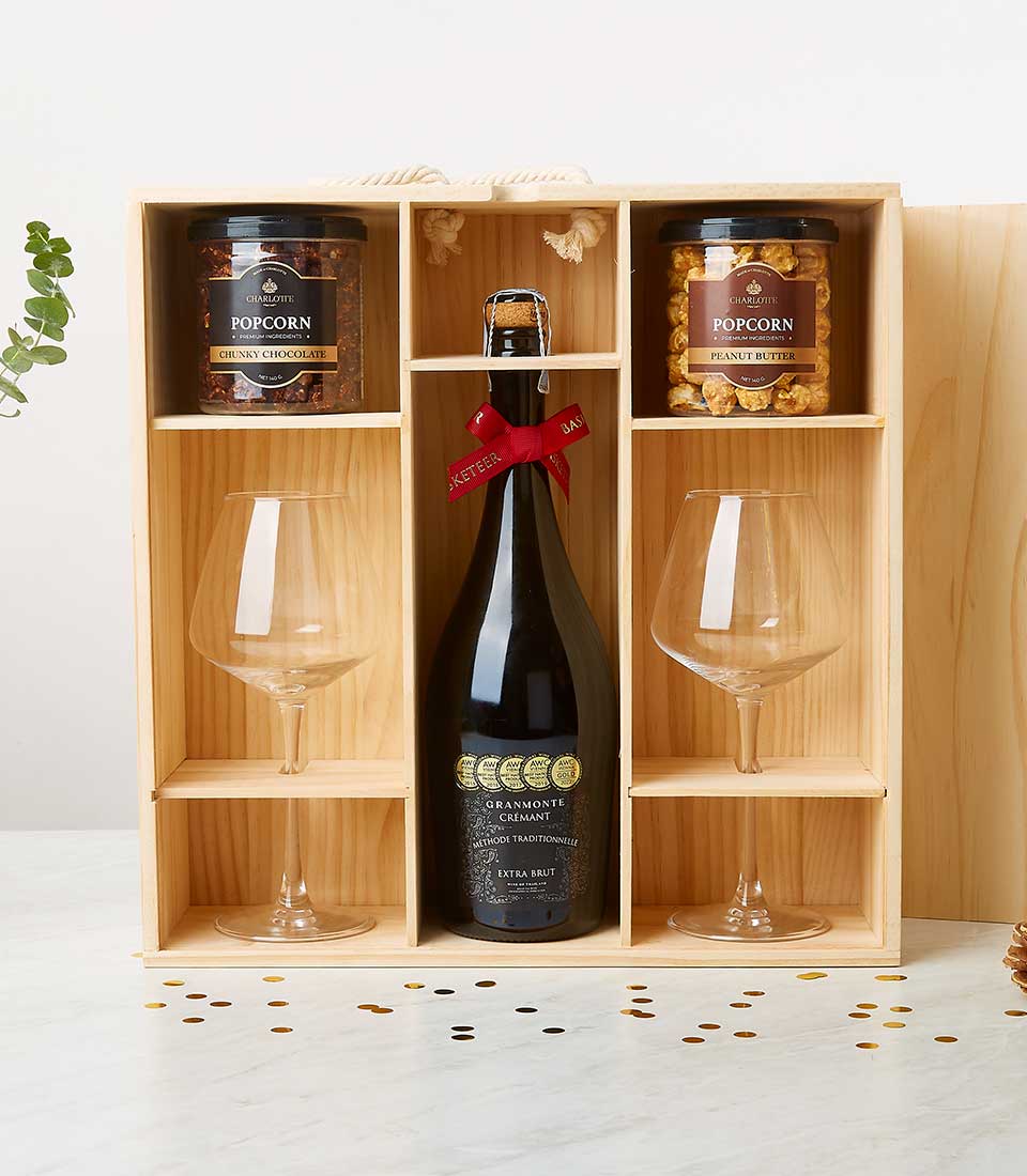 An opulent wooden gift box from Basketeer, adorned with a rich red ribbon, reveals a bottle of exquisite sparkling wine alongside two elegantly crafted wine glasses. Complementing the luxurious experience are two jars of gourmet flavored popcorn—one decadent chocolate and the other indulgent peanut butter. The scene is set on a light surface sprinkled with shimmering gold confetti, inviting you to savor every moment of this lavish offering.
