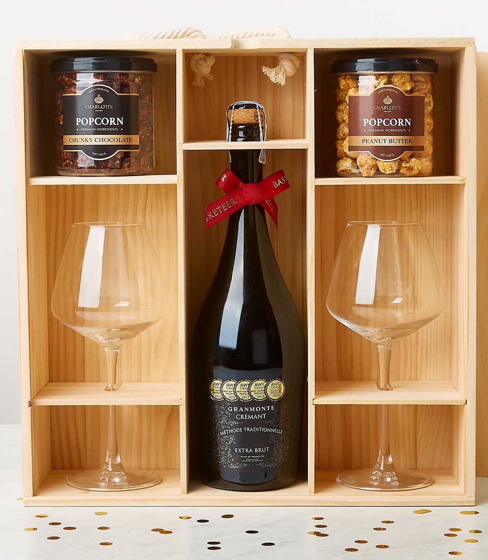 A luxurious Basketeer wooden gift box elegantly showcases a bottle of exquisite Gramonti crémant wine, accompanied by two pristine wine glasses ready for toasting. Nestled in their own compartments are two gourmet jars of popcorn, enticingly labeled 