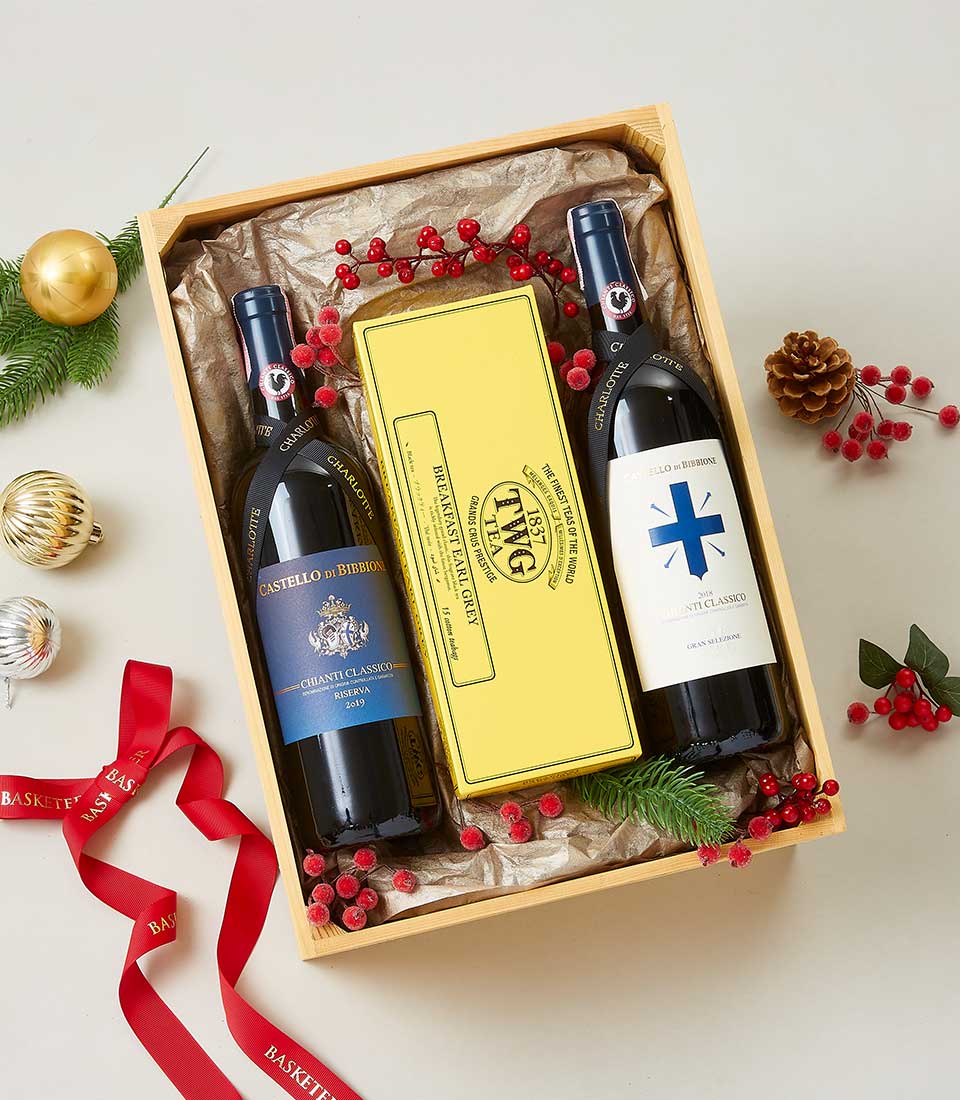 A lavish holiday gift box from Basketeer featuring two exquisite bottles of red wine, alongside a sophisticated yellow tea box. Adorned with rich red berries, an elegant pine cone, and lush evergreen sprigs, this sumptuous arrangement epitomizes festive luxury. The entire presentation is wrapped in a signature red ribbon bearing the Basketeer logo, making it an impeccable addition to the surrounding opulence of Christmas ornaments on a pristine white surface.