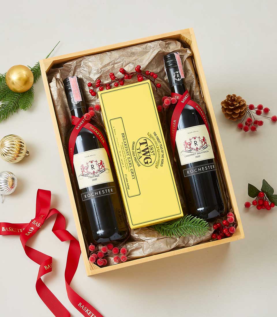 A luxurious and inviting gift box from Basketeer, adorned with an elegant red ribbon bearing the Basketeer logo, and filled with festive delights. Inside, two exquisite bottles of Rochester wine sit alongside a sumptuous, rectangular yellow box of gourmet cheese. The scene is beautifully decorated with vibrant red berries, lush green pine branches, and complemented by the presence of natural pinecones and gleaming gold Christmas ornaments.