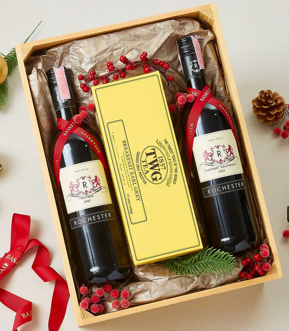 A luxurious gift box from Basketeer, exquisitely presenting two superb bottles of Rochester red wine and a distinguished rectangular yellow box of The Fine Cheese Co. Bath, England crackers. The beautifully arranged ensemble is adorned with festive red berries, elegant pine cones, and a sophisticated red ribbon bearing the Basketeer logo. Nestled on crinkled brown paper, this scene embodies opulence and warmth.