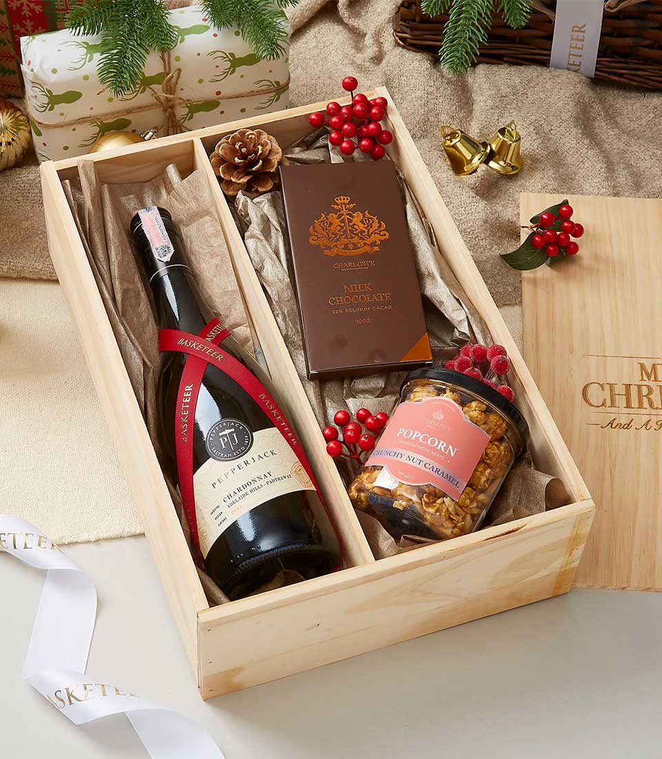 A luxurious wooden gift box from Basketeer showcases an exquisite arrangement designed to delight the senses. Nestled inside is an elegant bottle of red wine, a decadent bar of milk chocolate, and a gourmet jar of caramel popcorn. The box is adorned with festive holiday decorations, including bright holly berries and shimmering gold bells. A pristine white ribbon elegantly printed with 'Basketeer' ties the display together, adding an extra touch of sophistication and festivity to this perfect holiday gift.