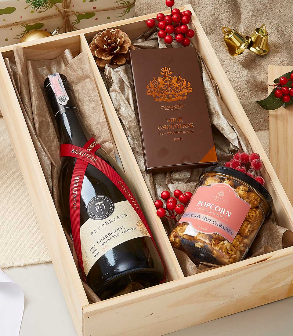A premium wooden gift box from Basketeer adorned with festive embellishments presents an elegant bottle of Pepperjack Shiraz, delicately tied with a luxurious red ribbon bearing the Basketeer logo. Nestled within the crinkled paper are indulgent treats—a creamy bar of milk chocolate and a jar of delectable crunchy nut caramel popcorn. Accentuating the scene are rustic pine cones and vibrant red berries, creating a captivating holiday ambiance.