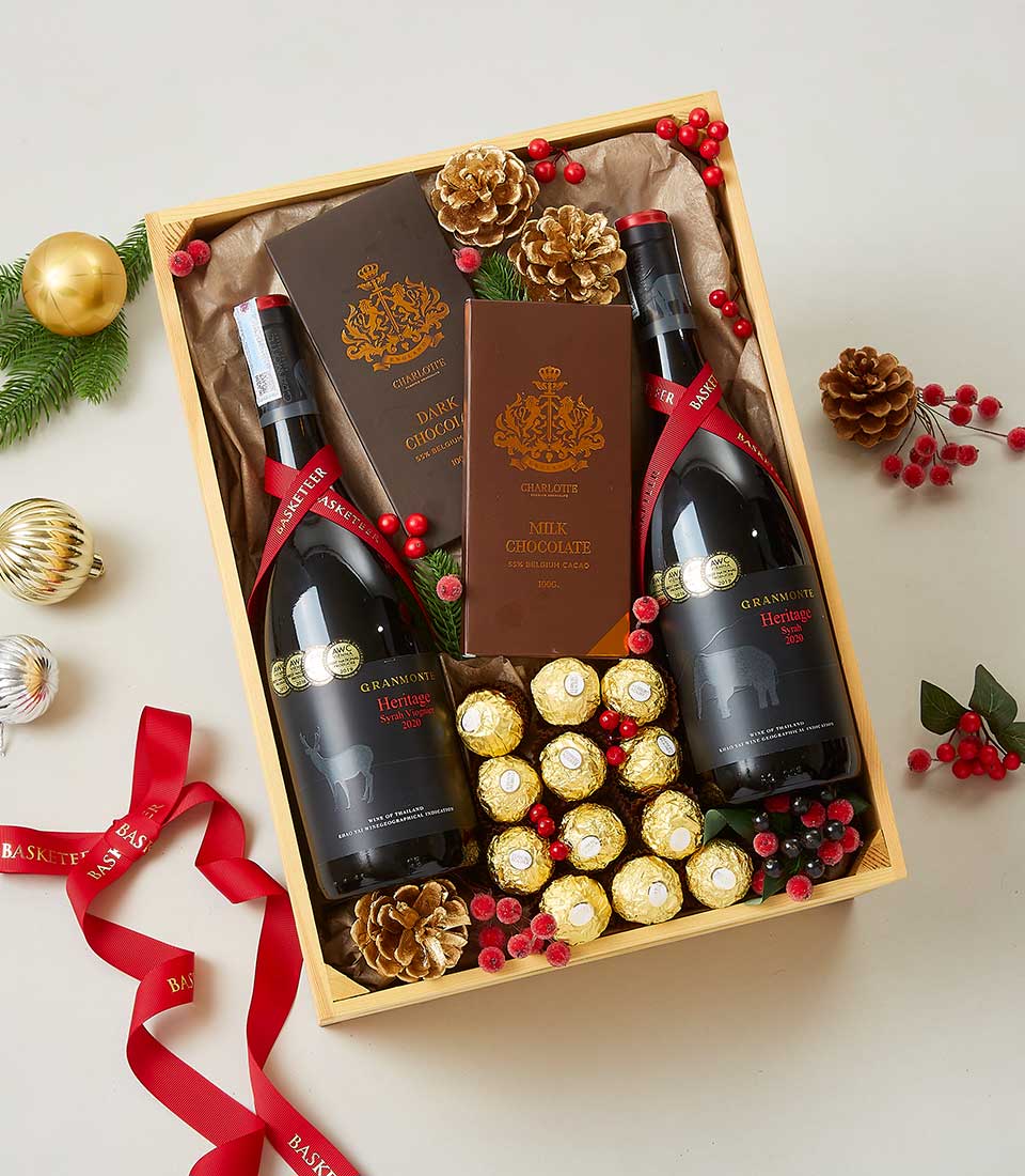 A luxurious gift basket by Basketeer, featuring two exquisite bottles of red wine and an assortment of decadent chocolates, including the iconic Ferrero Rocher. This elegant arrangement is adorned with rich red ribbons emblazoned with the Basketeer logo and festive touches such as glistening golden ornaments, charming pine cones, vibrant red berries, and a delicate sprig of green leaves. Luxuriate in the holiday spirit with this inviting scene that promises to delight and indulge.