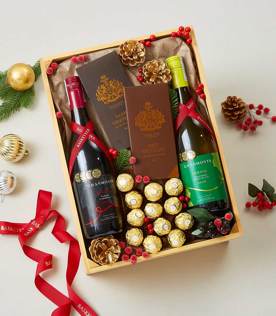A luxurious Basketeer festive gift box exquisitely presents two fine bottles of wine alongside an assortment of decadent chocolates and golden-wrapped truffles. The elegant box, adorned with the signature Basketeer logo, is beautifully decorated with rich red ribbons, pine cones, and holly berries. Surrounded by charming Christmas ornaments and lush greenery, this scene invites indulgence and festive cheer.