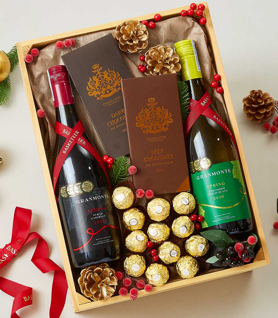 A luxurious wooden gift box from Basketeer showcases an exquisite arrangement designed to impress. Nestled inside are premium bottles of red and white wine, complemented by three decadent chocolate bars and an abundance of Ferrero Rocher chocolates. The presentation is beautifully enhanced with elegant pine cones, vibrant red berries, and a festive ribbon adorned with the Basketeer logo, making it a perfect holiday-themed indulgence.