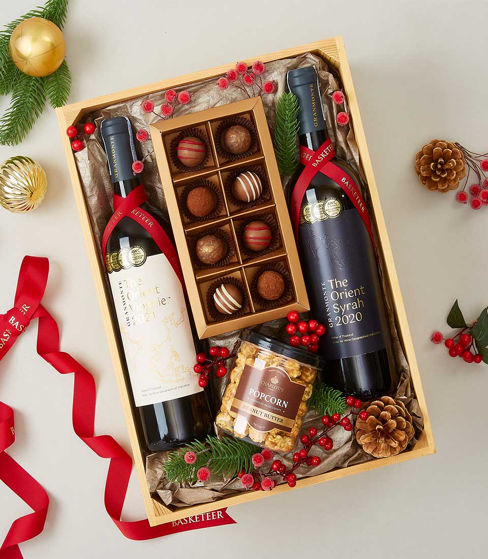 A luxurious wooden gift box featuring Basketeer branding showcases an inviting display of elegance and indulgence. Inside, you'll find two exquisite bottles of wine – a 2020 Sauvignon Blanc and a 2020 Syrah – perfectly nestled beside a jar of gourmet popcorn and an elegant box of assorted chocolates. The scene is made even more enchanting with festive decorations, including lavish red ribbons, charming pine cones, and vibrant red berries.