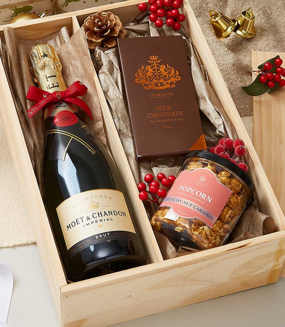 A luxurious wooden gift box elegantly showcases a bottle of Moët & Chandon champagne, paired with a decadent bar of Clarence Milk Chocolate and an exquisite jar of caramel-coated popcorn embellished with festive red berries. The Basketeer logo graces the ribbon on the box, symbolizing refined quality. Surrounding this sophisticated arrangement are decorative gold bells and vibrant red berry sprigs, creating an inviting and opulent scene.