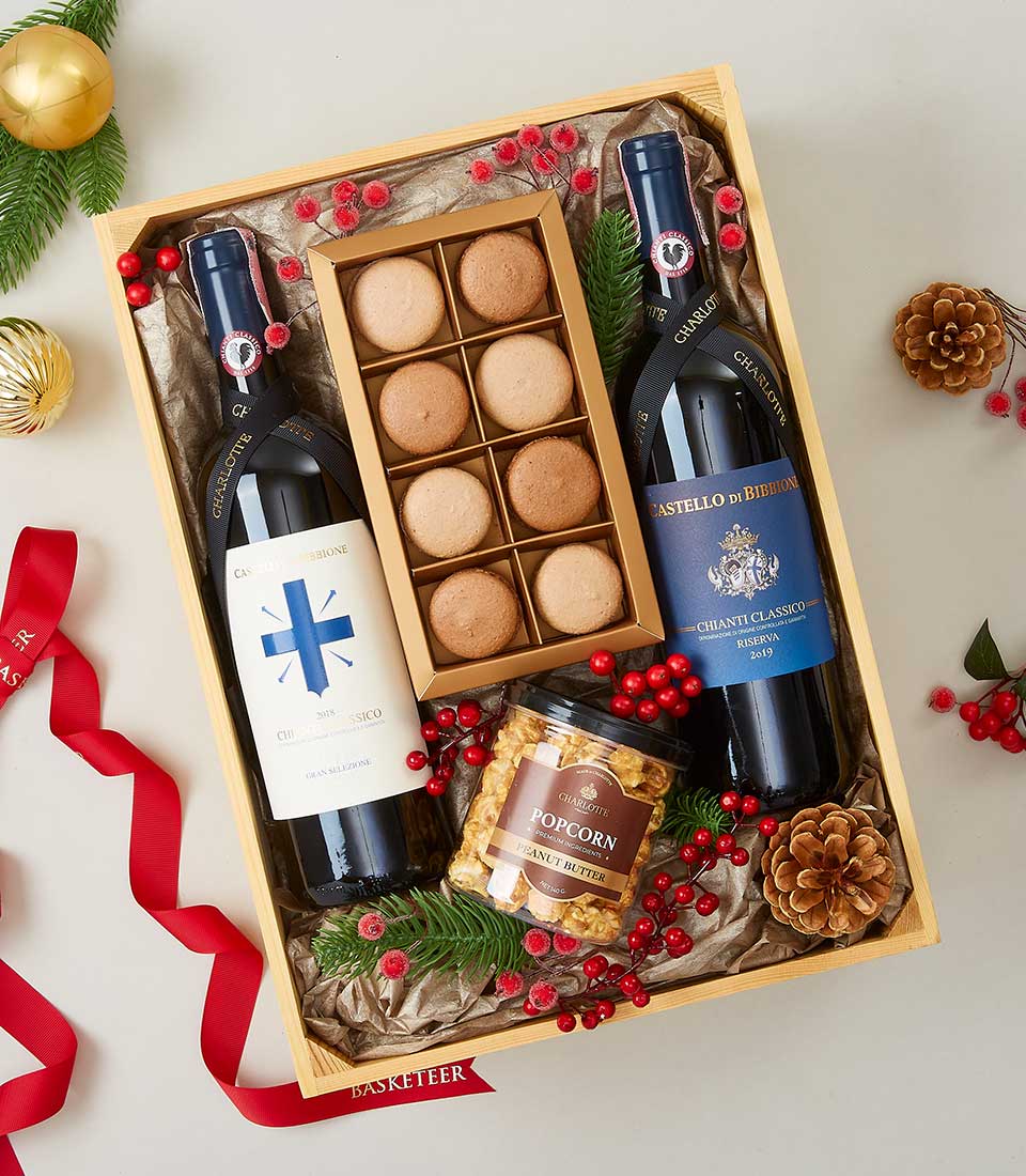 A luxurious wooden gift box from Basketeer, featuring two exquisite bottles of Chianti Classico wine. Nestled within are a gourmet container of Butter Toffee Popcorn and an elegant box of eight decadent macarons. The presentation is adorned with lush pine branches, charming pinecones, vibrant red berries, and a sophisticated red ribbon bearing the Basketeer logo—crafting an opulent and inviting holiday scene.