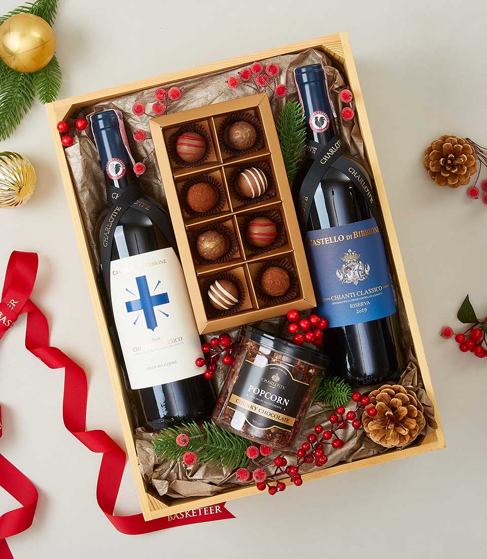 A lavish gift box from Basketeer, exquisitely presented with two elegant bottles of wine, an assortment of delectable chocolates, and a jar of indulgent caramel gourmet popcorn. The arrangement is tastefully adorned with charming pine cones, vibrant red berries, and lush green pine needles. A sophisticated red ribbon featuring the 