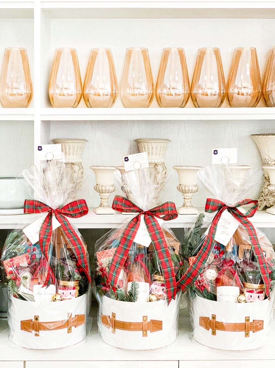 Three festive gift baskets, wrapped in clear plastic and adorned with red and green plaid ribbons, are displayed on a shelf. Each basket contains assorted holiday items. Behind them, a set of elegant glasses and decorative vases are neatly arranged.