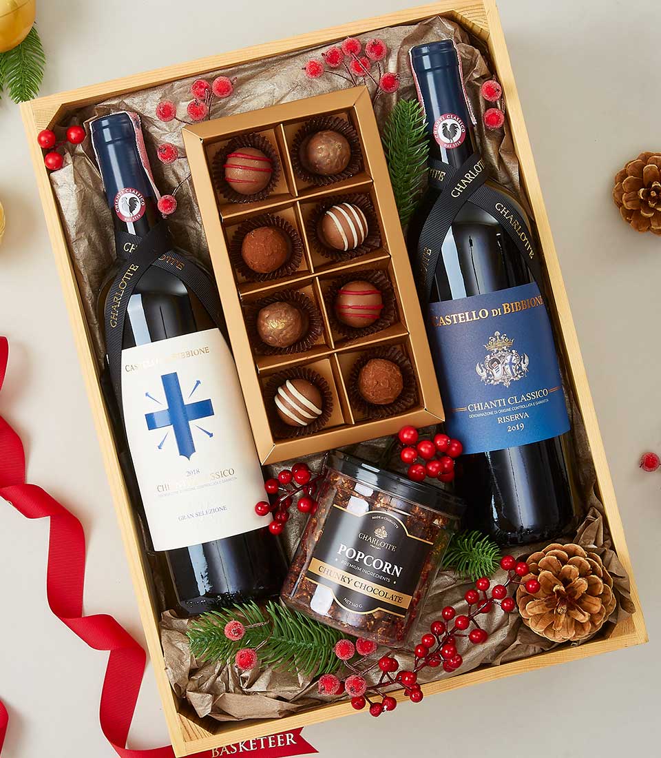 A luxurious Basketeer gift box exudes sophistication and festive cheer, beautifully adorned with our signature branding. Inside, you'll find two exquisite bottles of Chianti Classico wine nestled alongside a container of gourmet popcorn and an elegantly packaged assortment of decadent chocolates. The scene is made even more inviting with the addition of vibrant red berries, rustic pine cones, and a sumptuous red ribbon atop a bed of refined crinkled paper.