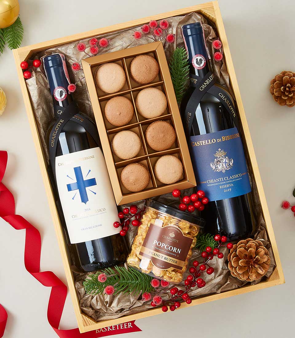 A luxurious wooden gift box from Basketeer showcases an assortment of indulgent delights: two exquisite bottles of fine wine, a pristine box containing nine delicate macarons, and a jar of sumptuous peanut butter popcorn. The scene is gracefully adorned with vibrant red berries, rustic pine cones, and lush greenery, evoking a sense of festive opulence. A sophisticated red ribbon featuring the Basketeer logo elegantly ties the presentation together, surrounded by artfully arranged pine sprigs that enhance its seasonal charm.