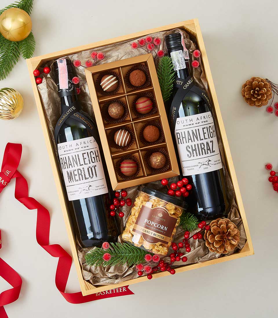 An exquisite wooden gift box from Basketeer, elegantly adorned with the brand's signature red ribbon. Inside, you'll discover two premium bottles of Rhanleigh Merlot and Rhanleigh Shiraz, each promising a rich and indulgent experience. Nestled alongside, a luxurious box of assorted chocolates tempts the senses while a jar of gourmet popcorn adds an artisanal touch. The scene is perfected with delicate accents of festive red berries, pinecones, and evergreen sprigs, inviting you to savor every moment of this opulent offering from Basketeer.