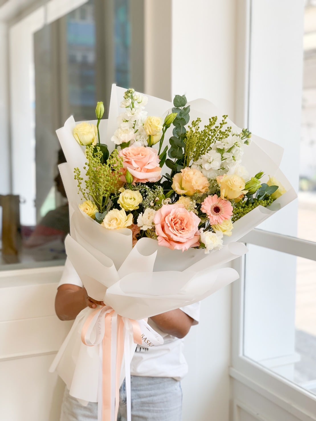 A person holding a large, elegant Radiance Bouquet wrapped in white paper. The bouquet includes dreamy blue roses, pink roses, white and yellow blooms, and greenery. The person is indoors, near a large window with natural light shining through.