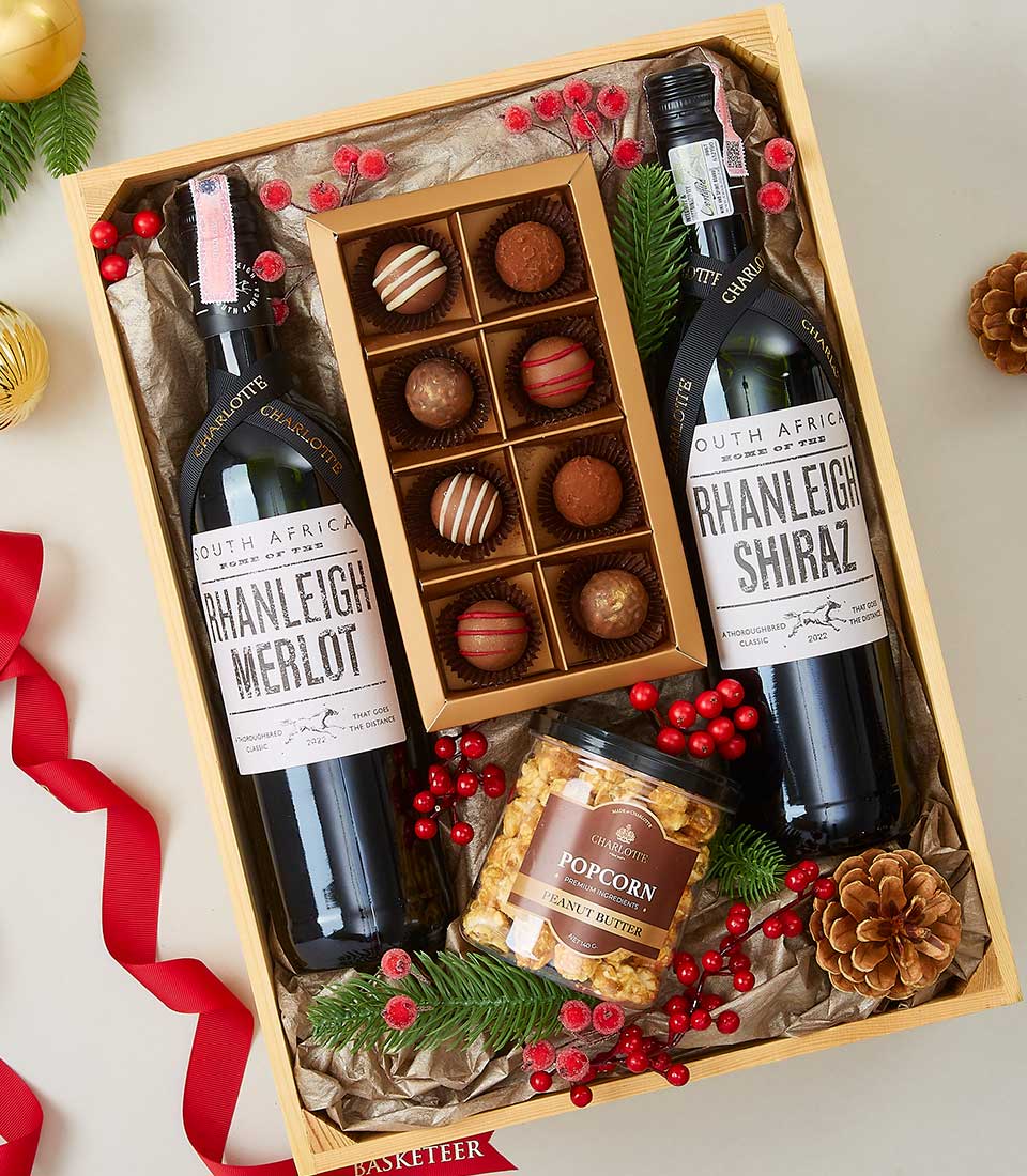 A lavish wooden gift box exudes opulence, featuring a distinguished bottle of Rhanleigh Merlot and a refined bottle of Rhanleigh Shiraz. Nestled alongside are a jar of gourmet caramel popcorn and an exquisite assortment of nine decadent chocolate truffles. Adorned with festive red berries, lush pine leaves, and an elegant red ribbon emblazoned with the Basketeer logo, this luxurious arrangement beckons celebration and indulgence.