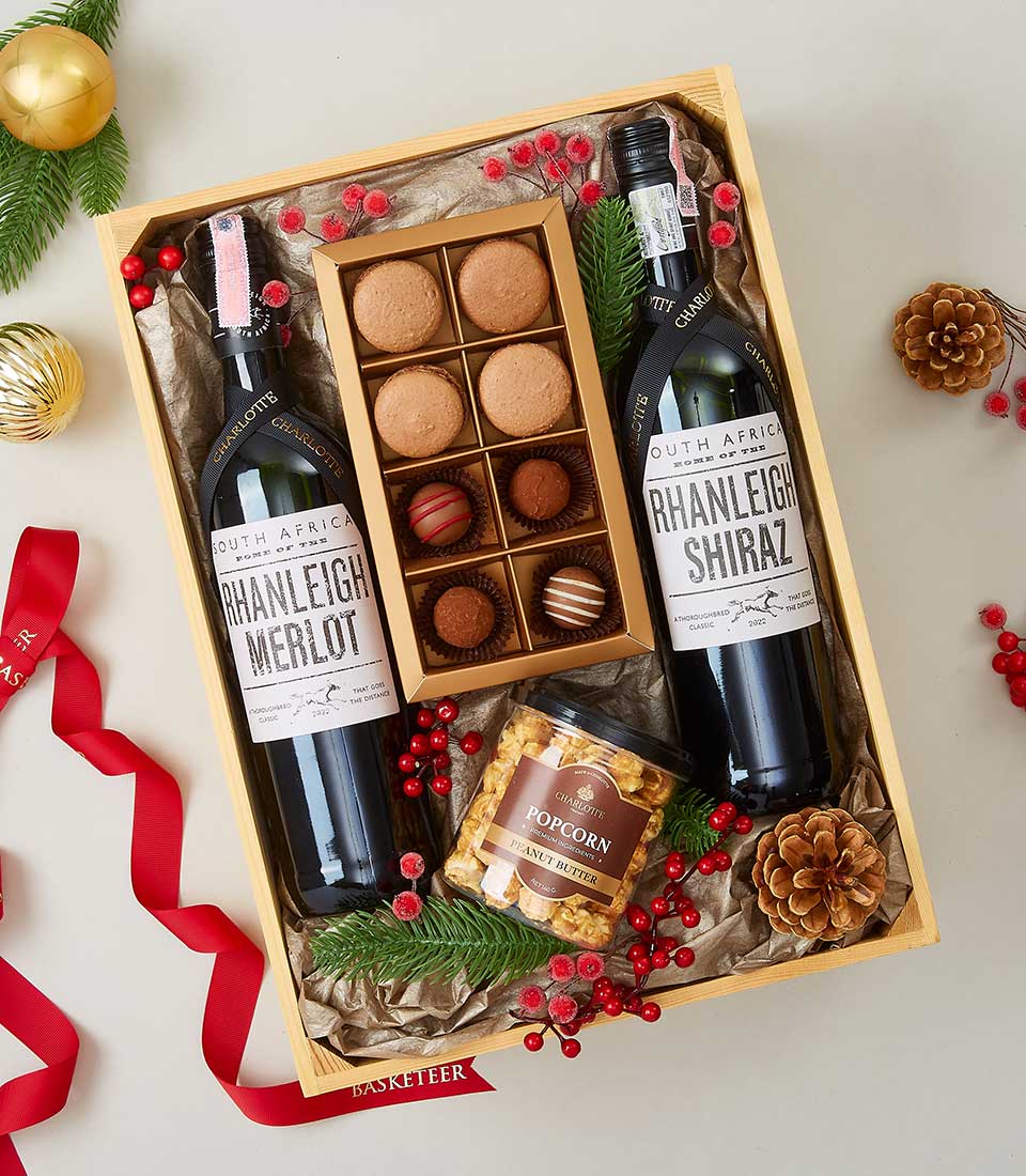 A luxurious and inviting gift box from Basketeer, exuding festive elegance. Nestled inside are two bottles of exquisite Rhanleigh wine (Merlot and Shiraz), accompanied by a gourmet tin of caramel popcorn and an array of decadent chocolates. The box is adorned with lush red berries, vibrant greenery, charming pinecones, and tied together with a satin ribbon featuring the Basketeer logo—creating a perfect blend of holiday cheer and sophistication.