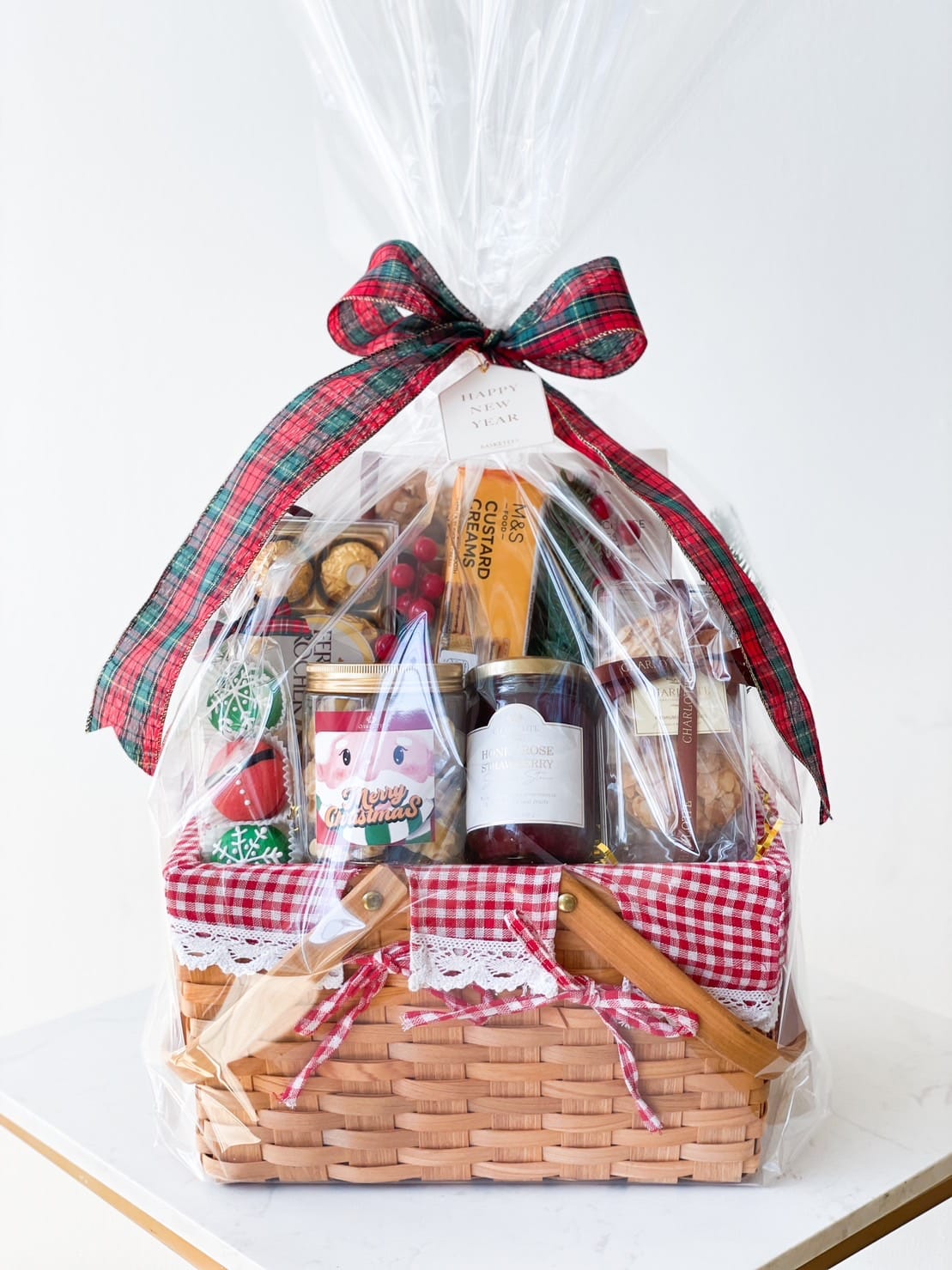 A gift basket wrapped in clear plastic and adorned with a red and green plaid ribbon. The basket contains various goodies, including a jar of fruit spread, a bottle of drink, assorted chocolates, and a pack of biscuits. The basket has a red gingham cloth liner with lace trim.