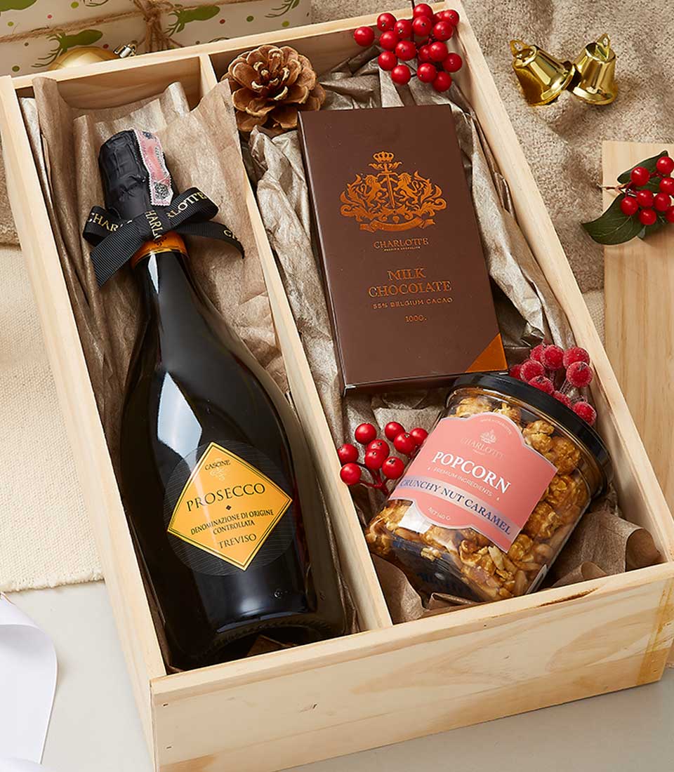 A luxurious wooden gift box from Basketeer showcasing a bottle of premium Prosecco, an indulgent bar of milk chocolate, and a gourmet container of toffee caramel popcorn. The scene is elegantly adorned with festive decorations, such as vibrant red berries, a charming pinecone, and tasteful gift wrap ribbons featuring the Basketeer logo, inviting you into a world of refined celebration and sumptuous delights.