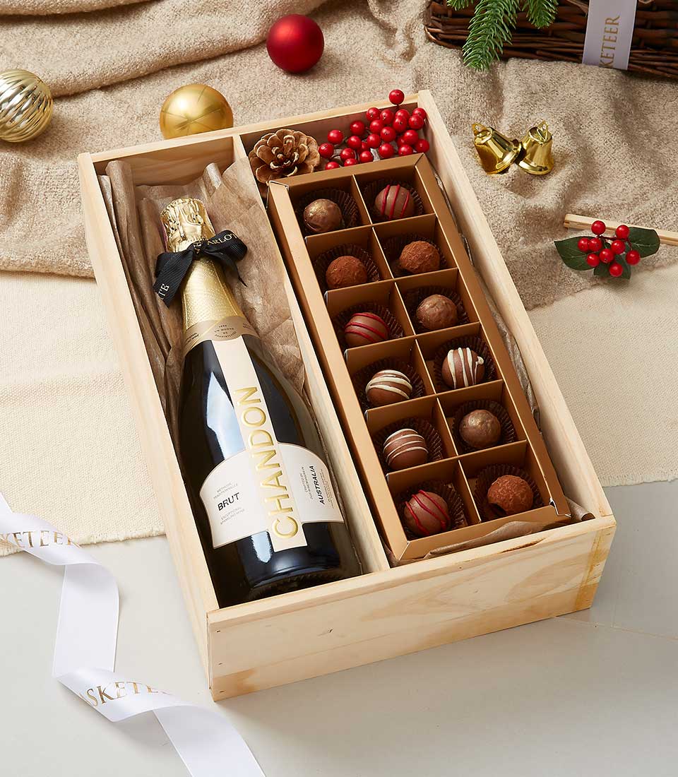 A luxurious wooden gift box from Basketeer features a premium bottle of Chandon Brut champagne elegantly placed on the left, while an assortment of decadent chocolate truffles awaits indulgence on the right. The exquisite presentation is enhanced by festive Christmas decorations, including shimmering ornaments, vibrant holly berries, rustic pinecones, and elegant bows. A stylish ribbon showcasing the distinguished Basketeer logo completes this opulent offering.
