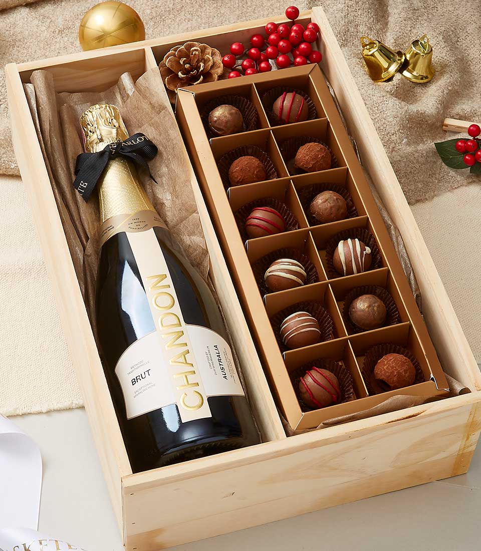 A luxurious wooden gift box from Basketeer showcases an exquisite bottle of Chandon Brut champagne accompanied by 16 decadent assorted chocolate truffles. The elegant presentation is enhanced with a festive pine cone and vibrant sprigs of red berries, exuding holiday charm. Nearby, shimmering golden bells and tasteful holiday decorations rest on an inviting textured beige cloth, completing the scene with a touch of opulence. The Basketeer logo subtly adorns the ribbon and box, symbolizing sophistication and elegance.