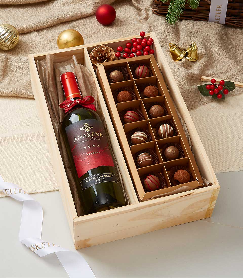 A lavish Basketeer wooden gift box set, artfully arranged with a bottle of Anakena Sauvignon Blanc wine on the left and an exquisite assortment of fine chocolates displayed in elegant rows on the right. The scene is adorned with festive Christmas ornaments, a textured burlap cloth, and a delicately draped ribbon showcasing the iconic 