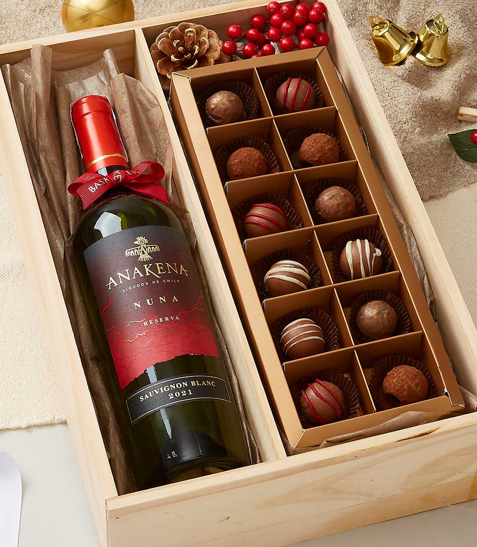 An exquisitely curated Basketeer gift box showcasing a distinguished bottle of Anakena Sauvignon Blanc wine, elegantly paired with a selection of 16 decadent chocolate truffles, arranged in an inviting grid. The box is adorned with festive embellishments including a rustic pine cone, vibrant red berries, and opulent golden bells, evoking the enchanting spirit of the holiday season. A luxurious ribbon branded with the Basketeer logo adds the perfect finishing touch to this sophisticated ensemble.