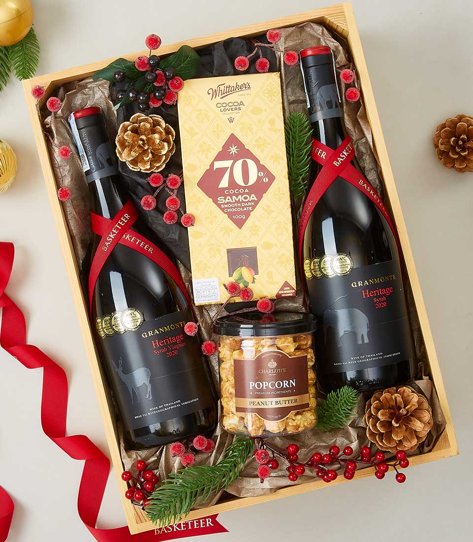 An exquisite holiday gift basket brimming with luxury awaits. Featuring two bottles of the revered Granmonte Heritage Syrah wine, this basket promises an experience of unparalleled sophistication. Indulge in the rich decadence of Whitaker's 70% Cocoa Samoa dark chocolate and savor the delightful crunch of a jar of gourmet peanut butter popcorn. Elegantly adorned with lush red ribbons, charming pinecones, and festive greenery, each detail is thoughtfully designed to elevate your gifting experience. The Basketeer branding on a ribbon or box adds an extra touch of prestige to this opulent presentation.