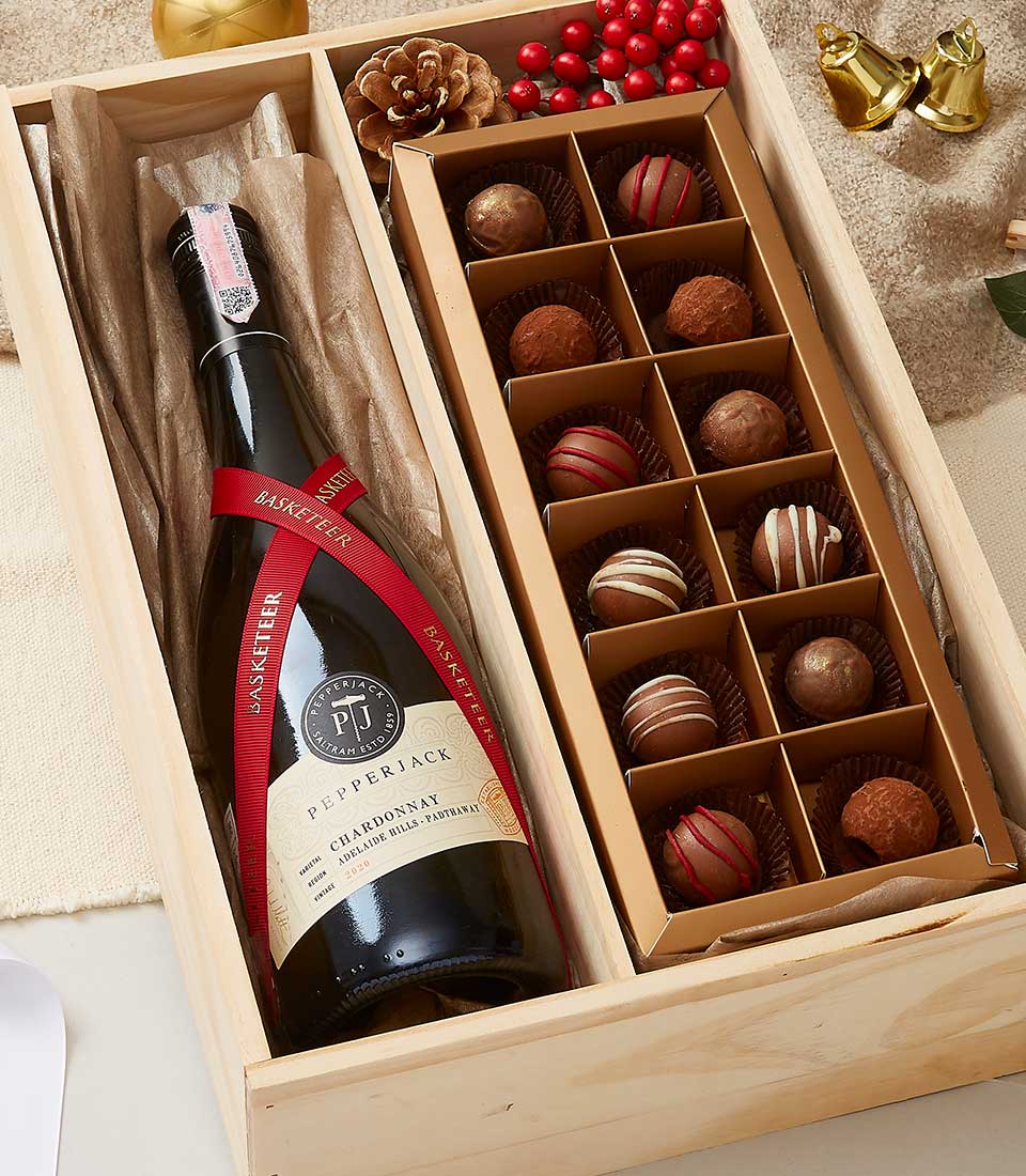 A lavish gift box from Basketeer showcases an exquisite bottle of Pepperjack Chardonnay, elegantly nestled in rich brown packing paper on the left side. To the right, a delectable selection of 12 assorted chocolate truffles is meticulously arranged in two perfect rows. The presentation is further enhanced with decorative pinecones, vibrant red berries, and festive adornments, creating an inviting holiday ambiance. A ribbon adorned with the Basketeer logo completes this luxurious ensemble.
