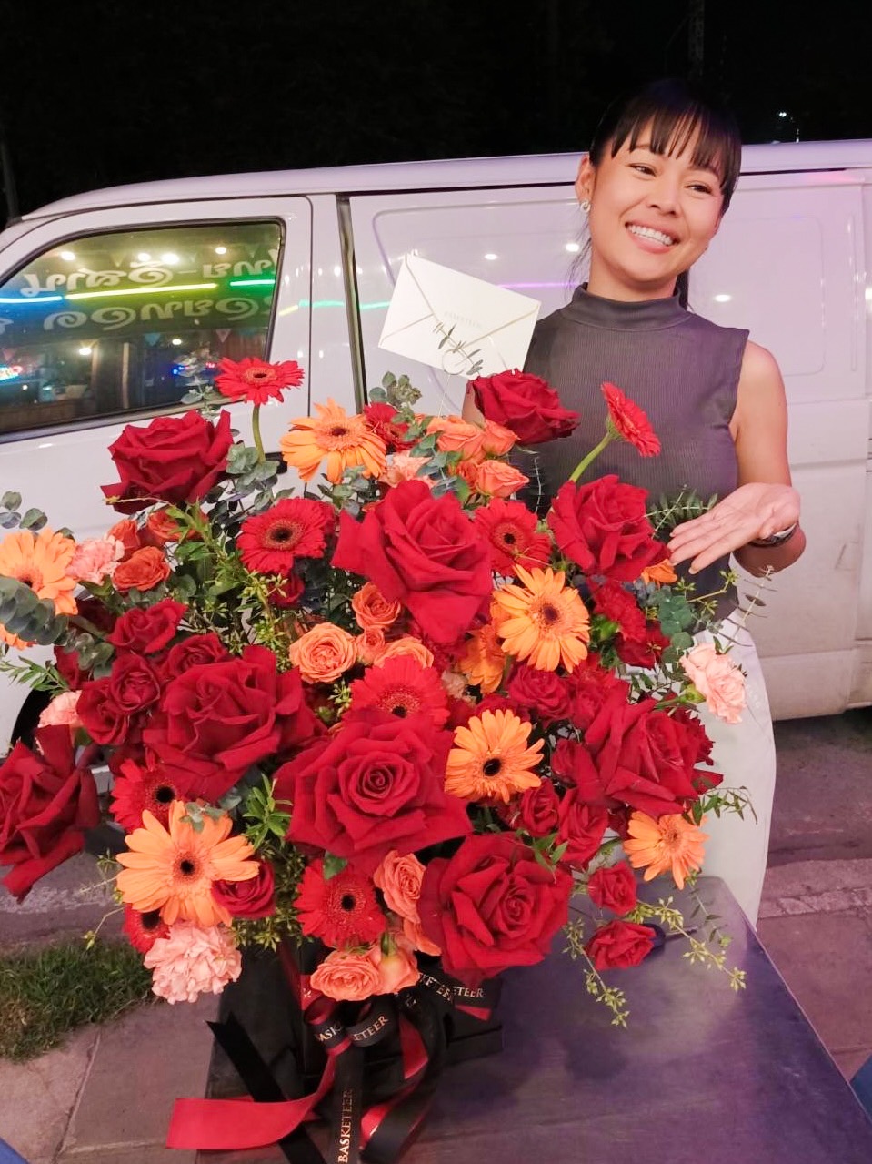 A woman dressed in a sleeveless grey top stands next to a large, vibrant bouquet of scarlet blooms, including red roses and orange flowers. She is smiling and holding a handwritten note. The backdrop features a white van and colorful neon lights, adding to the scene's romantic ambiance.