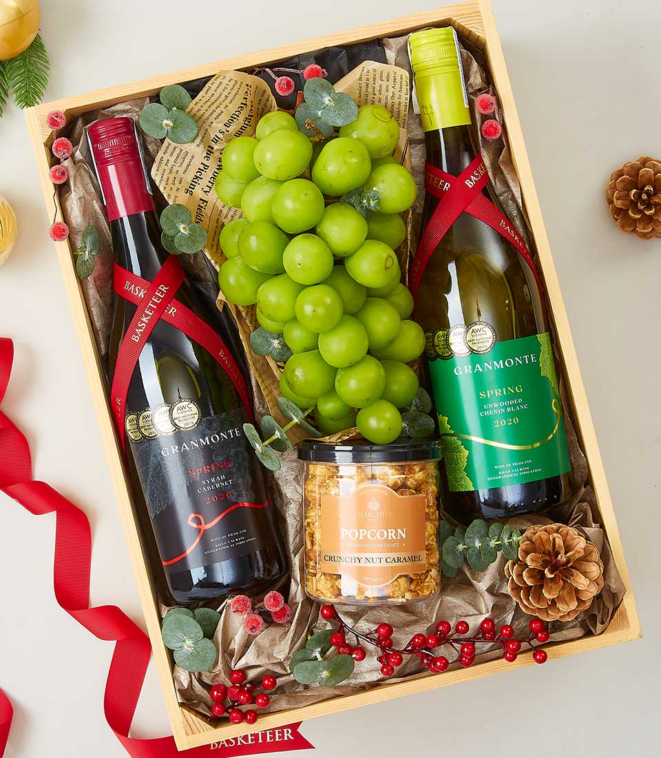 A lavish gift box by Basketeer showcases an exquisite collection: a bottle each of rich red and crisp white wine, nestled alongside a gourmet jar of crunchy nut caramel popcorn. A fresh, vibrant bunch of green grapes adds a touch of elegance. Complemented by seasonal pine cones, crimson red berries, and lush green decorative leaves, the scene is artfully adorned with luxurious red ribbons bearing the distinguished Basketeer logo.