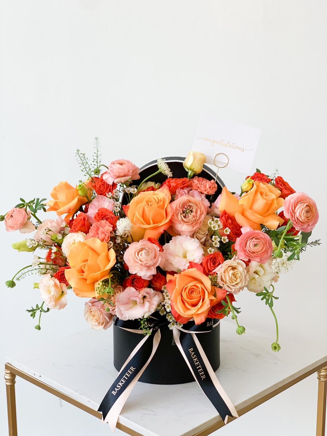 A black hatbox filled with a colorful arrangement of flowers, including orange roses, pink ranunculuses, white daisies, and green foliage. The box has white ribbons and a card that reads 