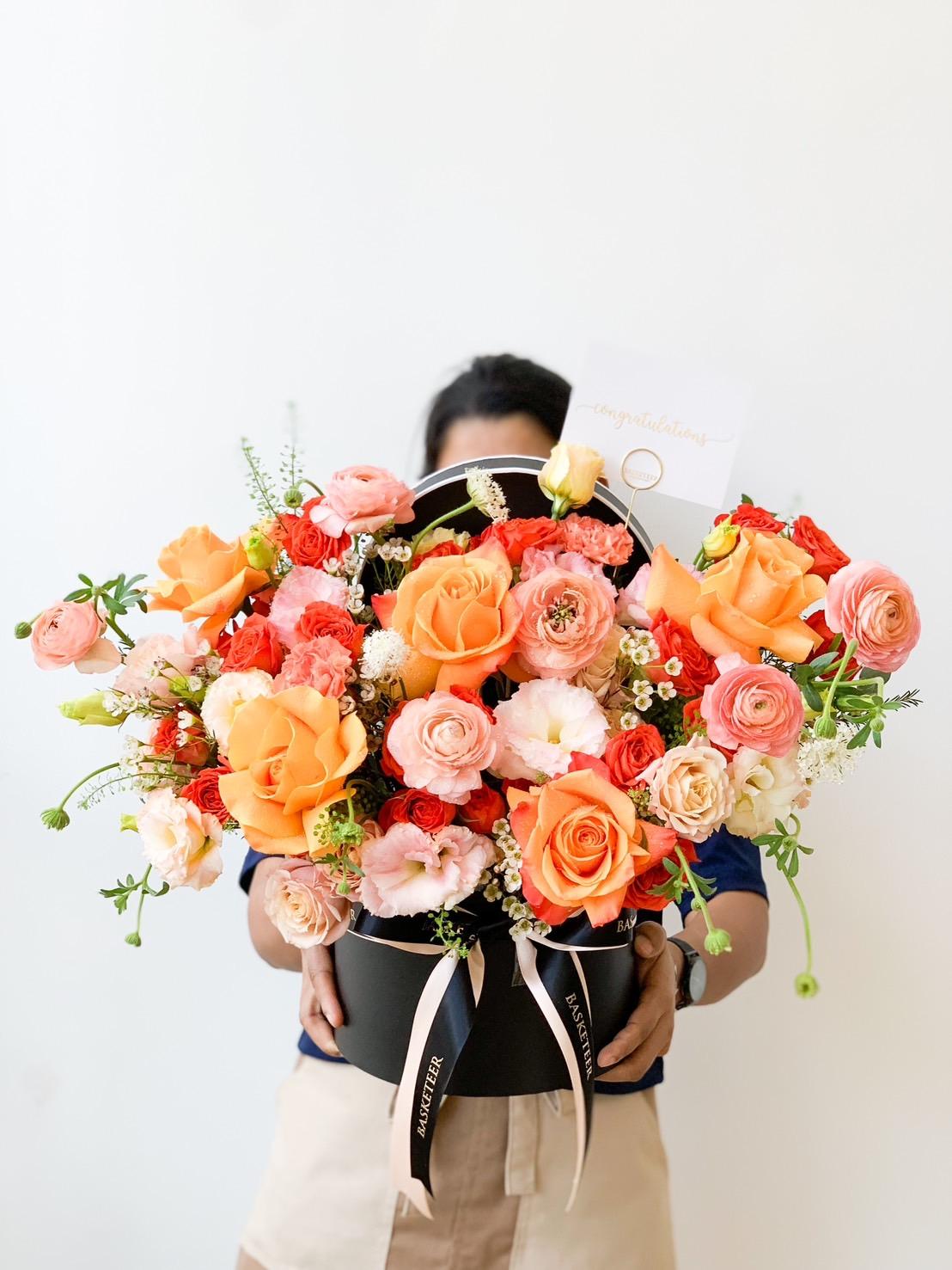 A person holds a large bouquet of vibrant orange, pink, and red flowers arranged in a circular black container. The bouquet is adorned with a ribbon and a small card that says 