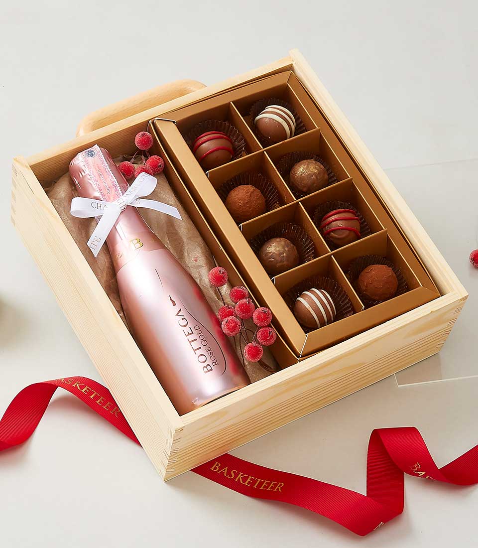 A luxurious wooden gift box elegantly displays a pink bottle of Bottega sparkling wine alongside an exquisite selection of assorted chocolates nestled within a sophisticated brown tray. The presentation is adorned with vibrant red berries and an elegant white ribbon, complemented by the distinguished presence of a red ribbon showcasing the Basketeer logo. This opulent scene invites indulgence and refinement, embodying the exceptional quality and style synonymous with the Basketeer brand.