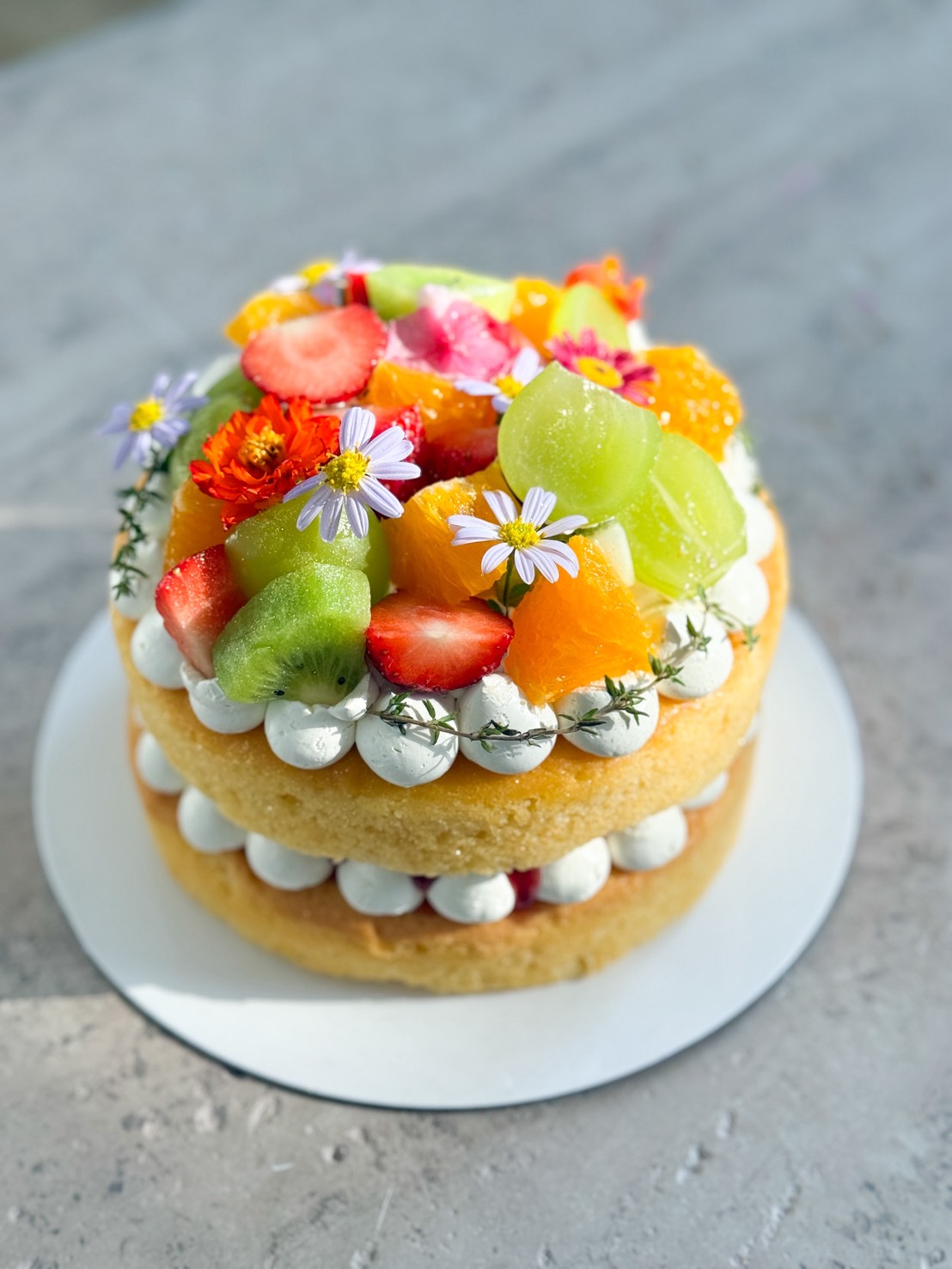 A Summery Strawberry Cream Cake, this two-layer sponge is topped with whipped cream, sliced strawberries, kiwi, orange segments, and edible flowers on a round white plate against a grey background.