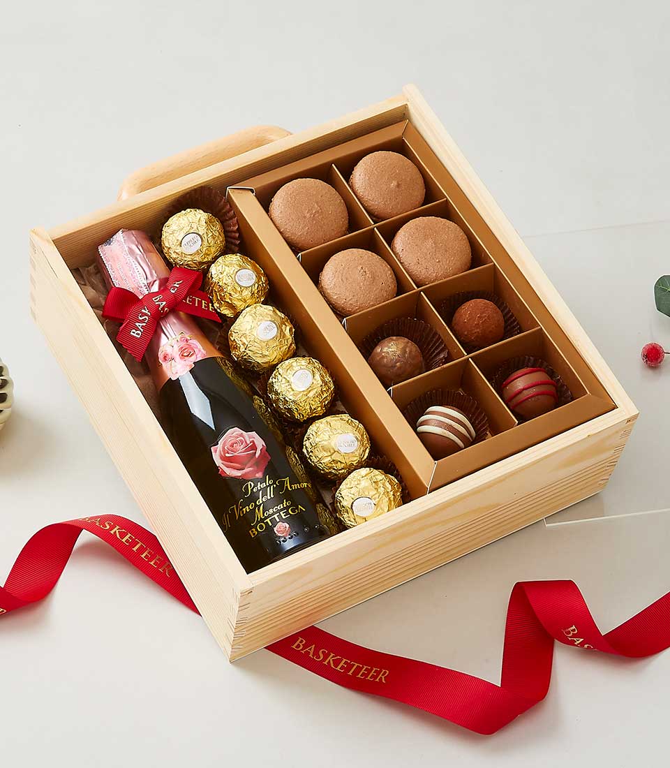 A luxurious wooden gift box curated by Basketeer, featuring a bottle of exquisite sparkling wine, an array of decadent chocolates, vibrant macarons, and opulent truffles. Each treat is meticulously organized within the box's elegant compartments. The presentation is perfectly accentuated with a lavish red ribbon proudly displaying the 