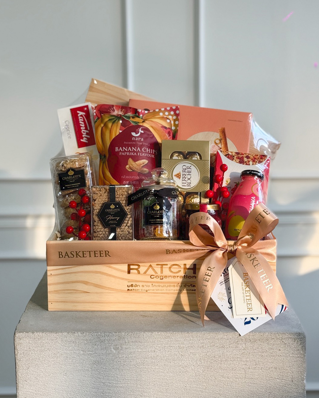 The Gourmet Woodland Delights Hamper is a wooden gift hamper filled with gourmet treats like banana chips, chocolates, nuts, jam, biscuits, and a bottled drink, all wrapped in clear plastic and adorned with a bow and gift tags.