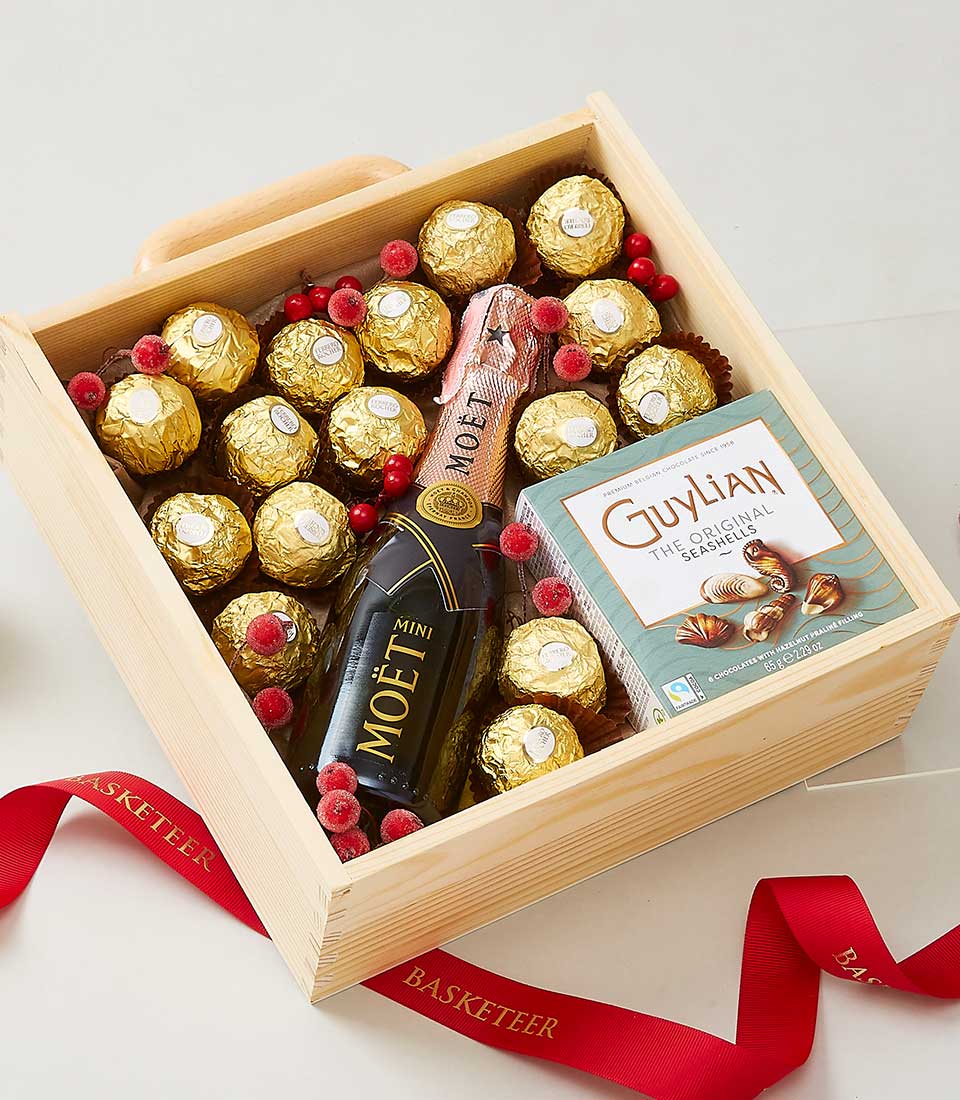 A lavish wooden gift box showcasing a mini bottle of Moët & Chandon champagne, nestled alongside an exquisite box of Guylian chocolates and several Ferrero Rocher chocolates, each meticulously wrapped in gold foil. Red berries add a festive touch, all elegantly tied together with a luxurious red ribbon flaunting the distinguished 