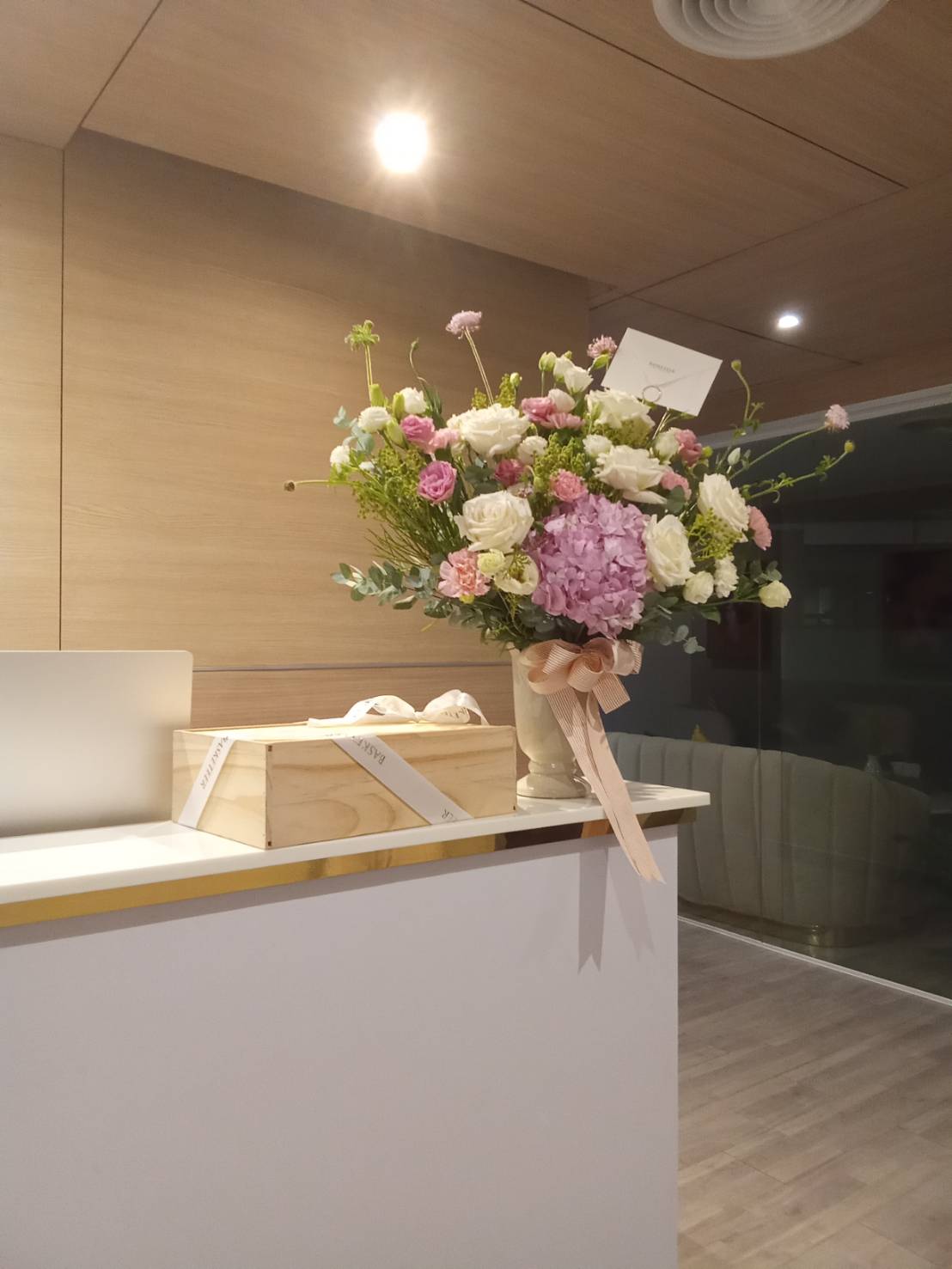 A bouquet of various flowers in pastel shades, arranged in a vase tied with a pink ribbon, is placed on a white countertop. Next to it is a wooden box containing two baked items wrapped with a ribbon. The background features a modern, wood-paneled interior.
