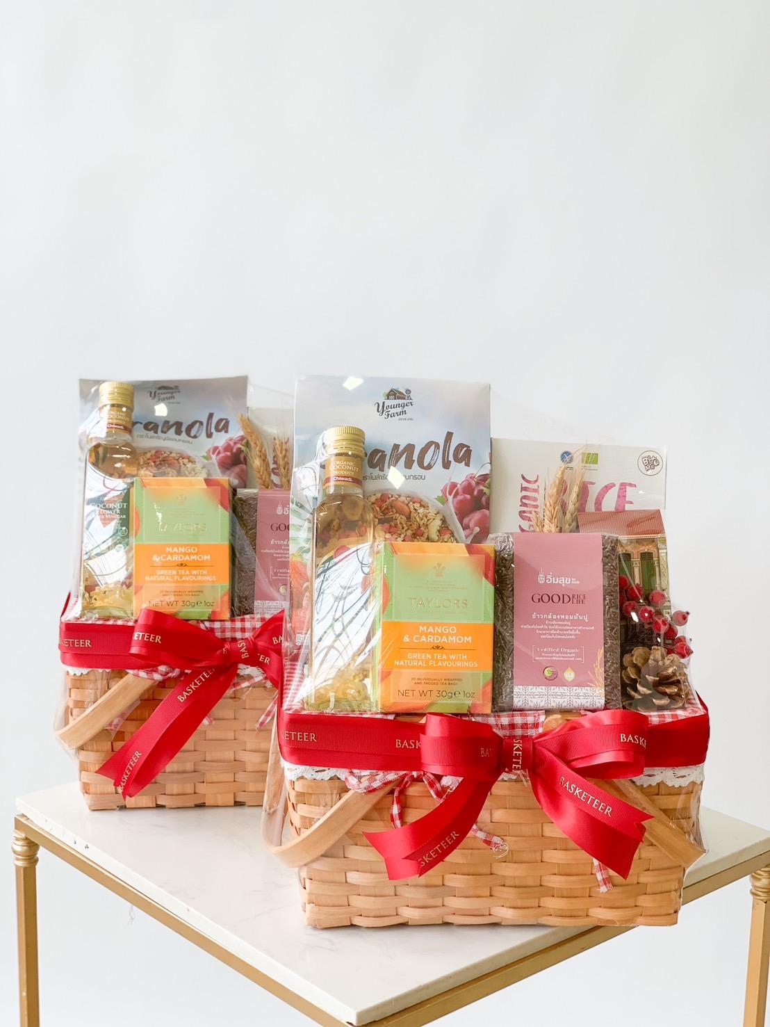 Three gift baskets are displayed on a table, each containing items like granola, pasta, and other gourmet food products. The baskets are decorated with red ribbons, adding a festive touch to the presentation.