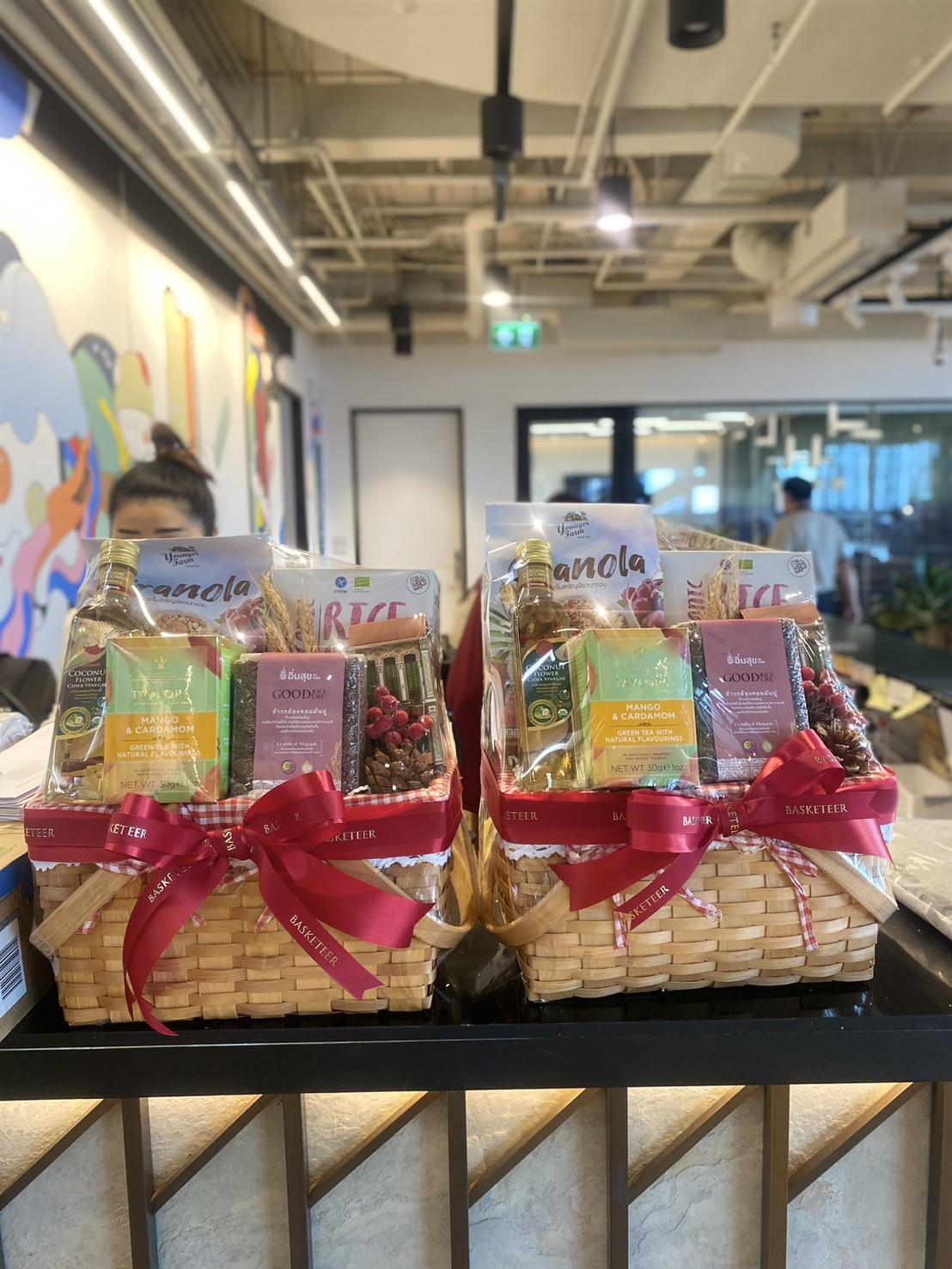 Two wicker gift baskets are displayed on a counter in an office setting. Each basket is wrapped with a red ribbon and filled with various snacks, including granola packets, chocolate boxes, and bottles of oil. People can be seen in the blurred background.