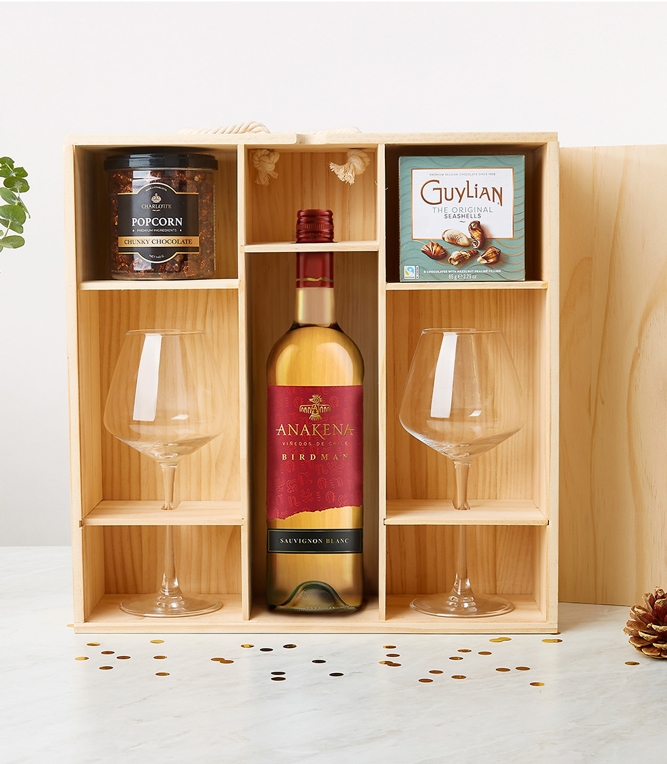 An elegant wooden gift box beautifully showcases a bottle of Los Vascos red wine adorned with a sumptuous red bow, accompanied by two pristine wine glasses. Nestled within are a jar of delectable Chunky Chocolate Popcorn and an exquisite box of Guylian Milk Chocolate Belgian truffles. The luxurious scene is further accentuated with scattered gold and black confetti on the white surface beneath, adding a touch of celebration. A ribbon inscribed with the Basketeer logo subtly hints at the curated opulence within this mesmerizing gift set.