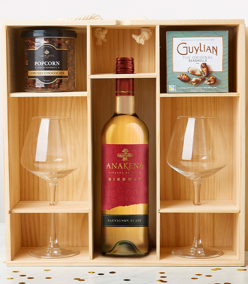 An elegant wooden gift box beautifully showcases a bottle of Los Vascos red wine adorned with a sumptuous red bow, accompanied by two pristine wine glasses. Nestled within are a jar of delectable Chunky Chocolate Popcorn and an exquisite box of Guylian Milk Chocolate Belgian truffles. The luxurious scene is further accentuated with scattered gold and black confetti on the white surface beneath, adding a touch of celebration. A ribbon inscribed with the Basketeer logo subtly hints at the curated opulence within this mesmerizing gift set.