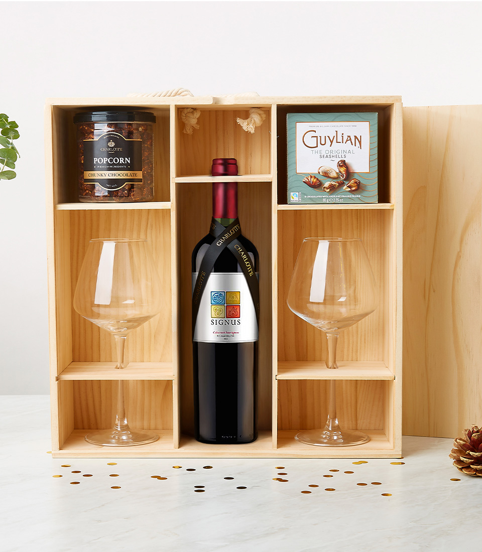 A luxurious wooden gift box from Basketeer elegantly showcases a bottle of exquisite Tempus Two wine, flanked by two crystal wine glasses. Nestled above the bottle, a jar of decadent organic chocolate-coated popcorn sits alongside a sophisticated box of Guylian Belgian chocolates. Delicate decorative confetti is artfully scattered below, adding an extra touch of celebration and opulence to the scene. The prestigious Basketeer logo graces a ribbon and box within the arrangement, seamlessly tying together this invitation to indulge in refined elegance.